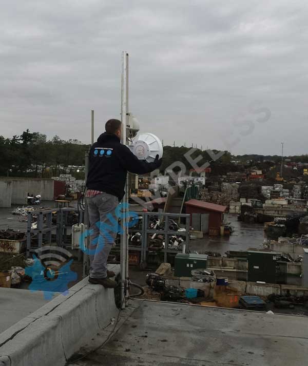 Installer on a rooftop aligning a white dish antenna on a pole above an outdoor industrial yard.