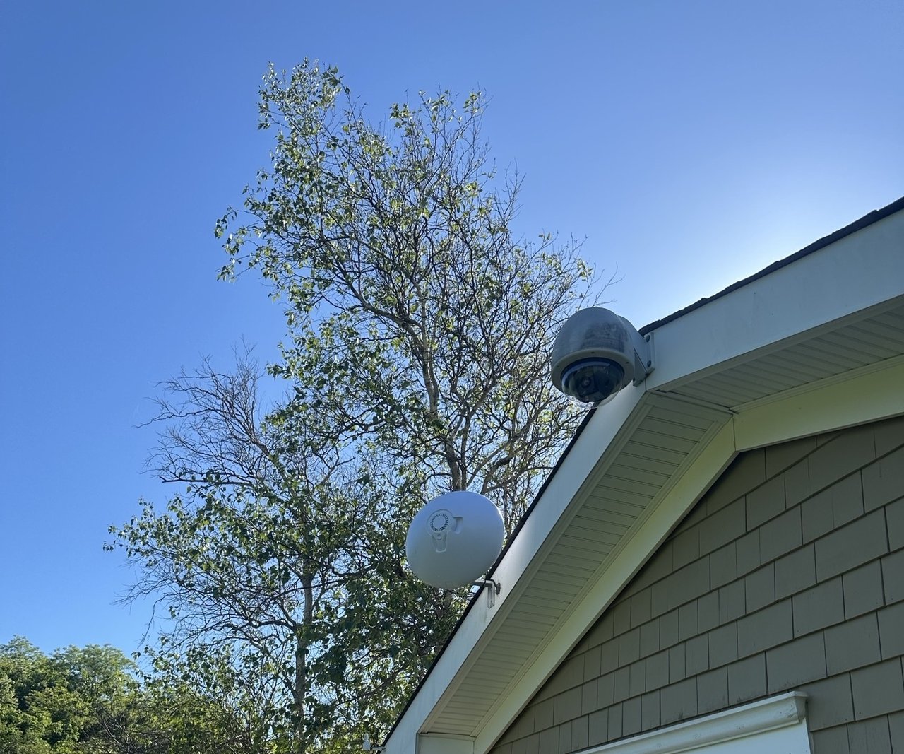 White circular wireless bridge and a dome camera mounted on a building soffit under a clear sky.