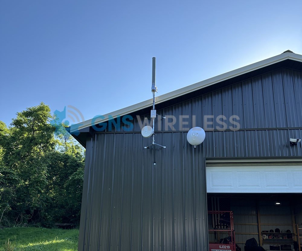 Outdoor antennas and dish radios mounted on a gray metal building beside trees and a clear sky.