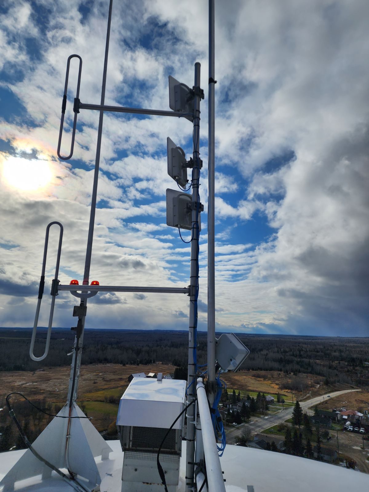 Antennas and cables on metal masts at the top of a high outdoor structure, with fields, trees, and a road below a cloudy sky.