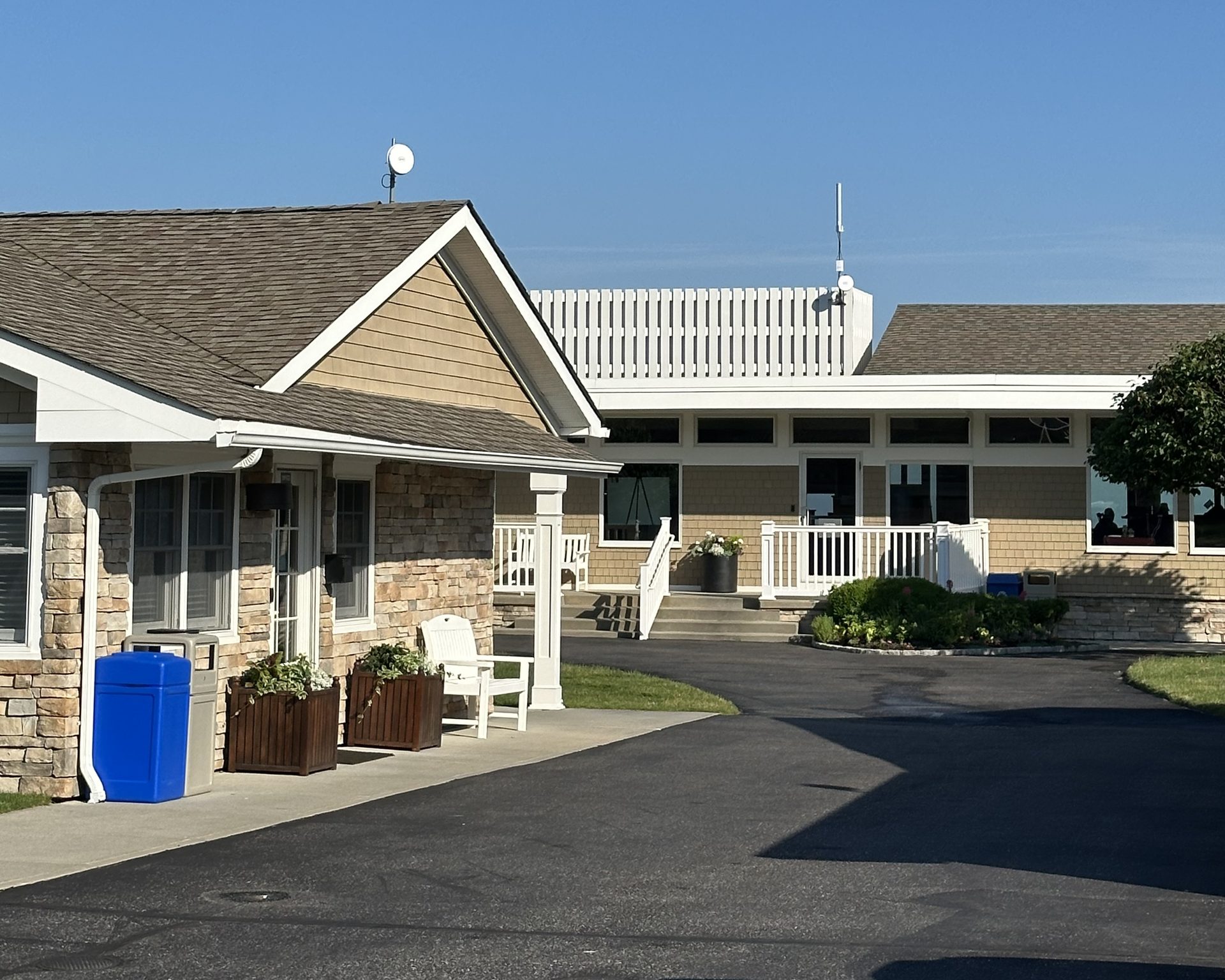 White circular dish antennas on two building roofs and a tall white vertical antenna, with tan siding, stone accents, and white railings under a clear blue sky.