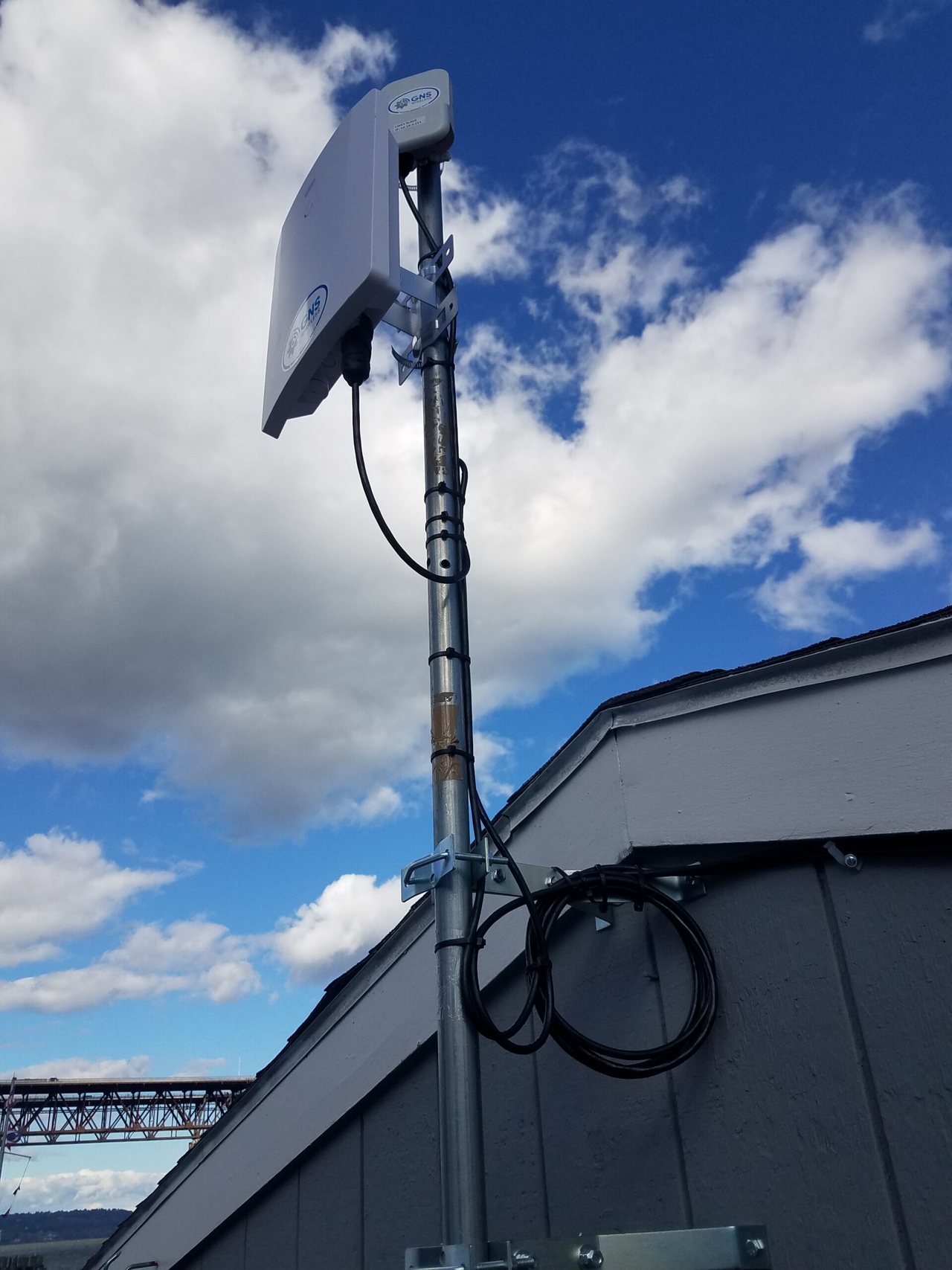 White rectangular outdoor antenna mounted high on a galvanized mast beside a building, with black cable secured down the mast.