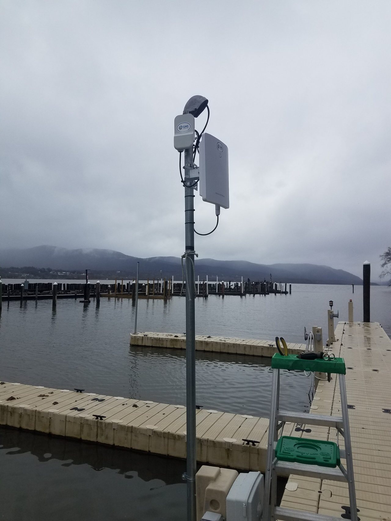 Galvanized pole on a floating marina dock with two white outdoor wireless units at the top, a grey junction box at the base, and a stepladder on the dock.