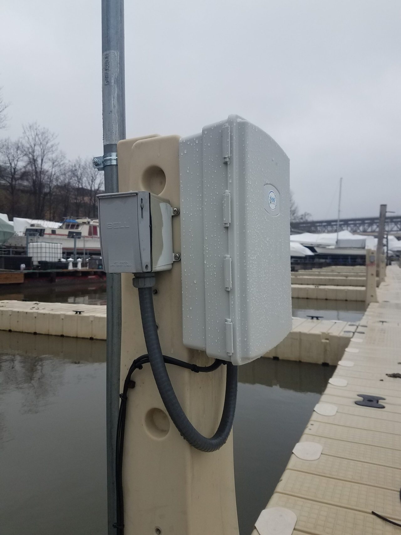 Weatherproof enclosure and outdoor electrical box mounted on a marina utility pedestal, wet with rain, with winter-covered boats in the background.