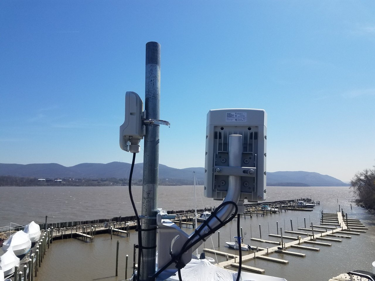 Outdoor wireless radios on a metal pole, with a smaller access point and a larger directional radio above a marina